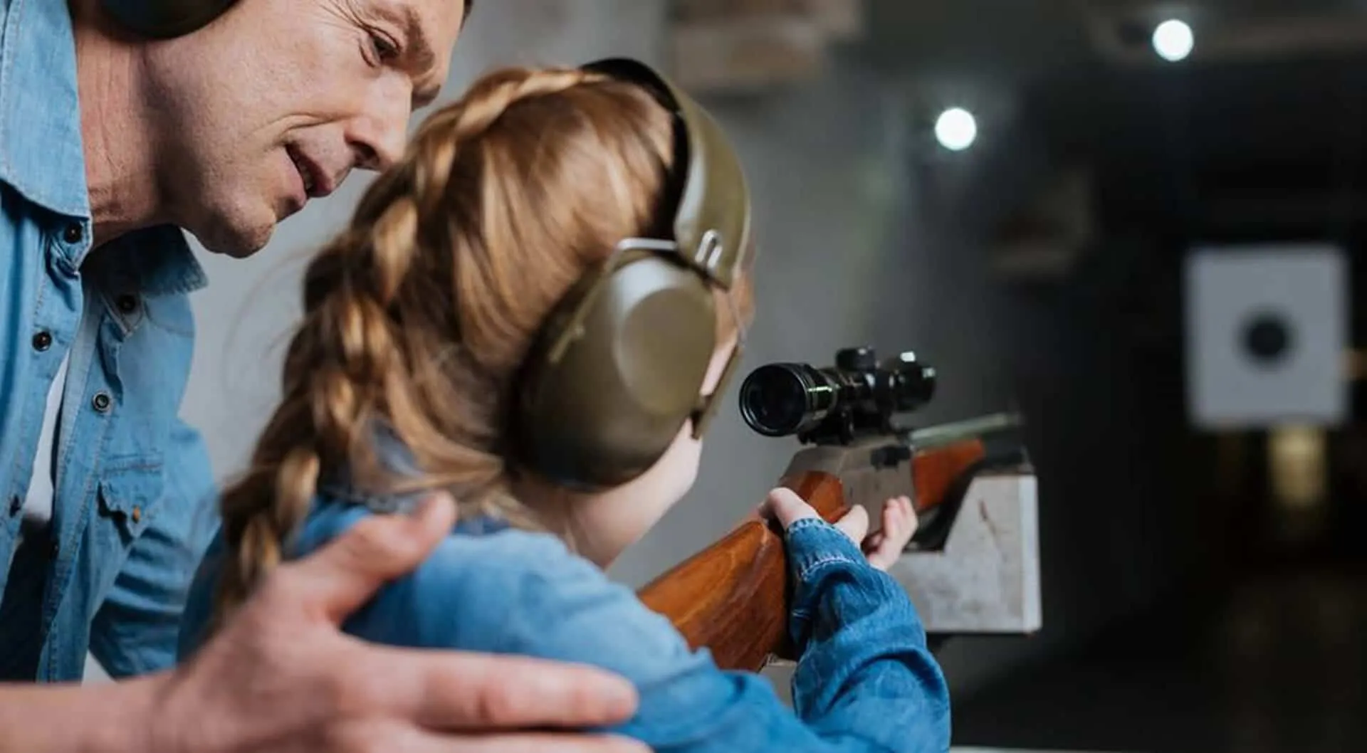 A father helping a daughter shoot a rifle at a gun range.